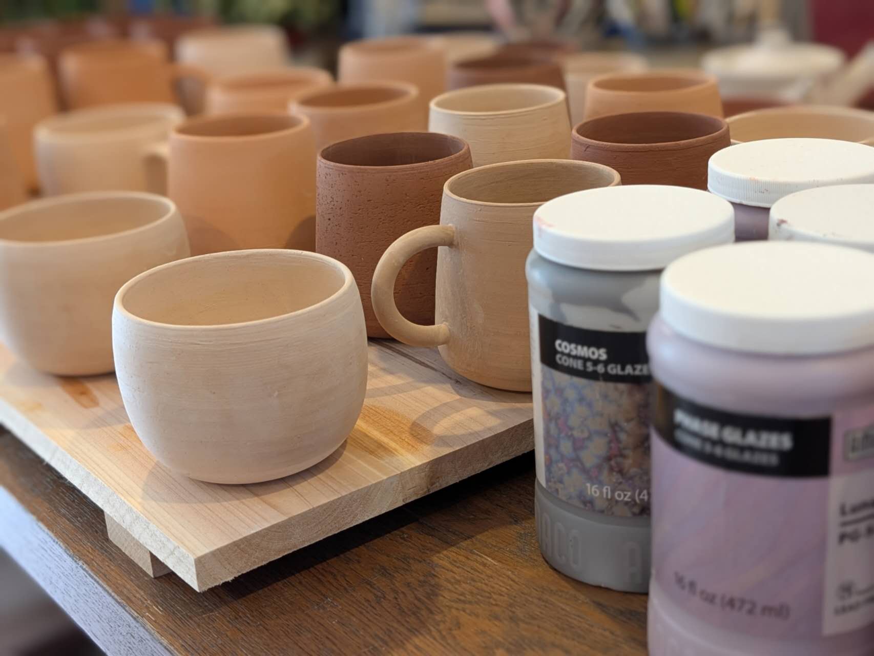 Clay mugs on a wooden board with glaze containers in the foreground